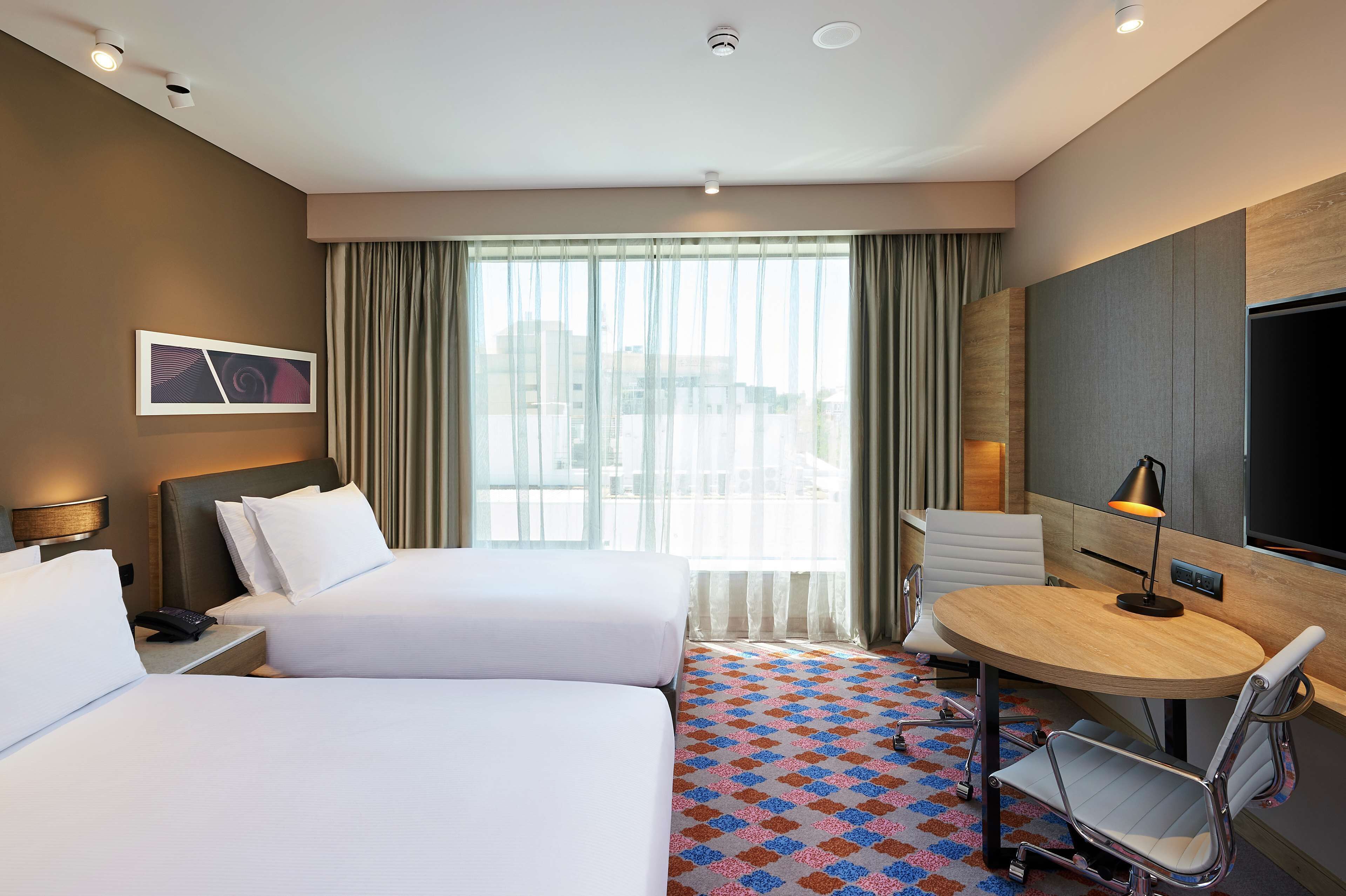 A hotel room with two beds, a desk, a chair, a TV, and a large window with curtains.