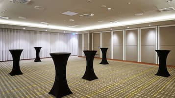 Sala de conferências com mesas pretas, carpete estampado e teto com iluminação embutida e ventilação.