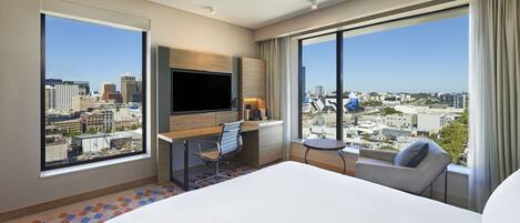 A hotel room with a large bed, a desk with a chair, a TV, and a view of the cityscape through large windows.