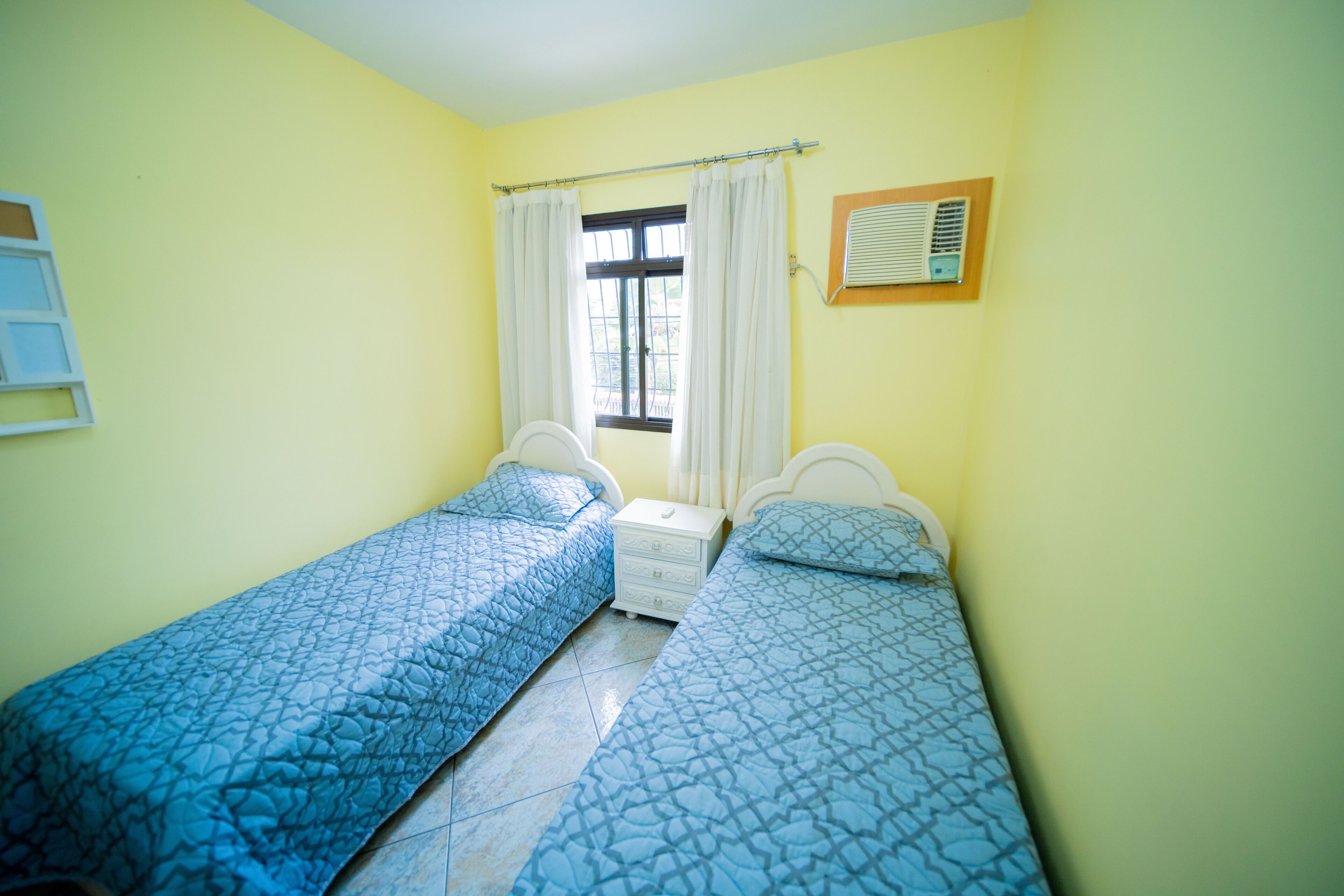 A room with two single beds, a window with curtains, a bedside table, and an air conditioning unit on the wall.