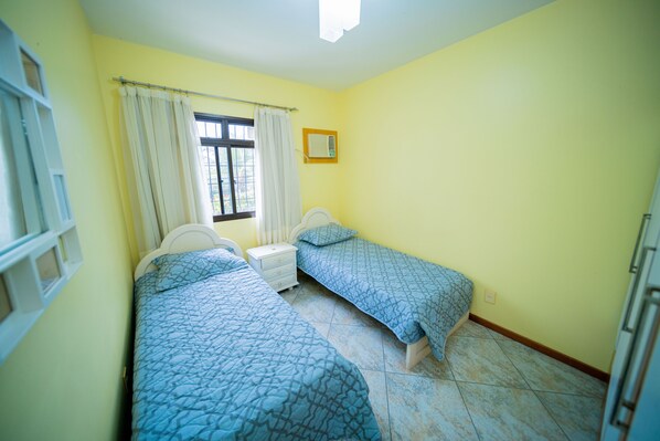Una camera da letto con due letti singoli, una finestra con tende, un comodino e un condizionatore d'aria a parete.