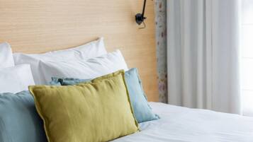 Cama bem arrumada com almofadas brancas e coloridas, cabeceira de madeira e luminária preta fixada na parede.