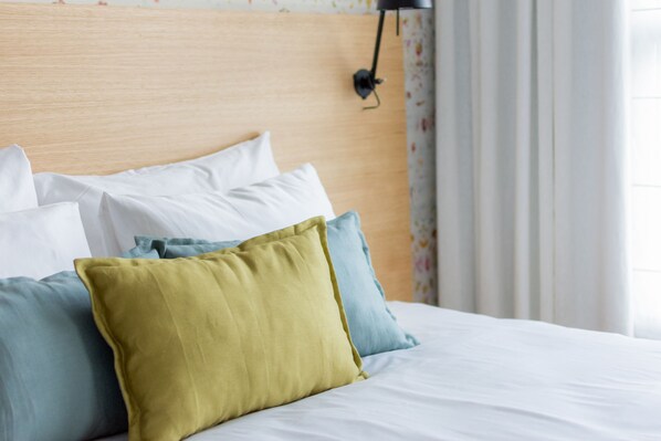 Una cama bien hecha con almohadas blancas y de color, un cabecero de madera y una lámpara negra fijada a la pared.