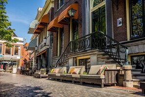 A row of outdoor seating with a stone balustrade, a brick building with large windows, and a clear blue sky. - Boutique Hotel Catshuis (Leeuwarden)