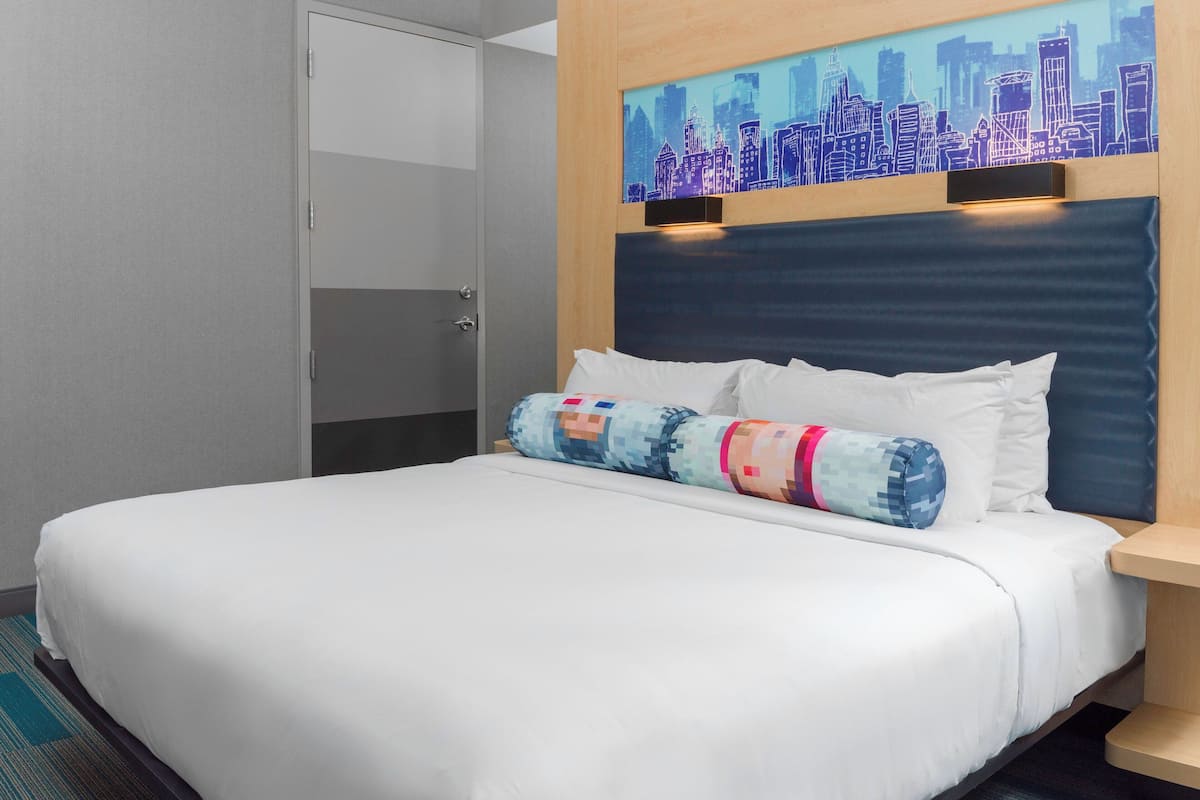 A hotel room with a bed, a cityscape artwork on the headboard, and a door to the left.