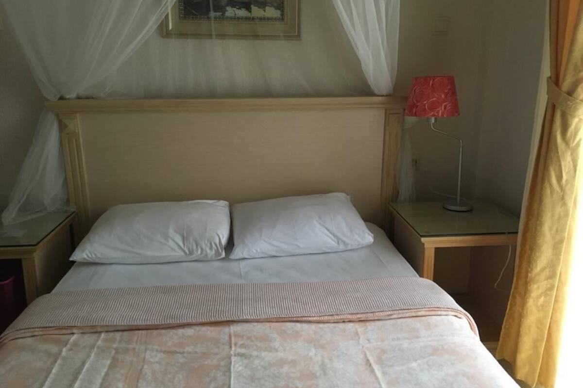 Un lit avec du linge de lit blanc, une tête de lit en bois, une table de chevet avec une lampe rouge et un tableau accroché au mur.
