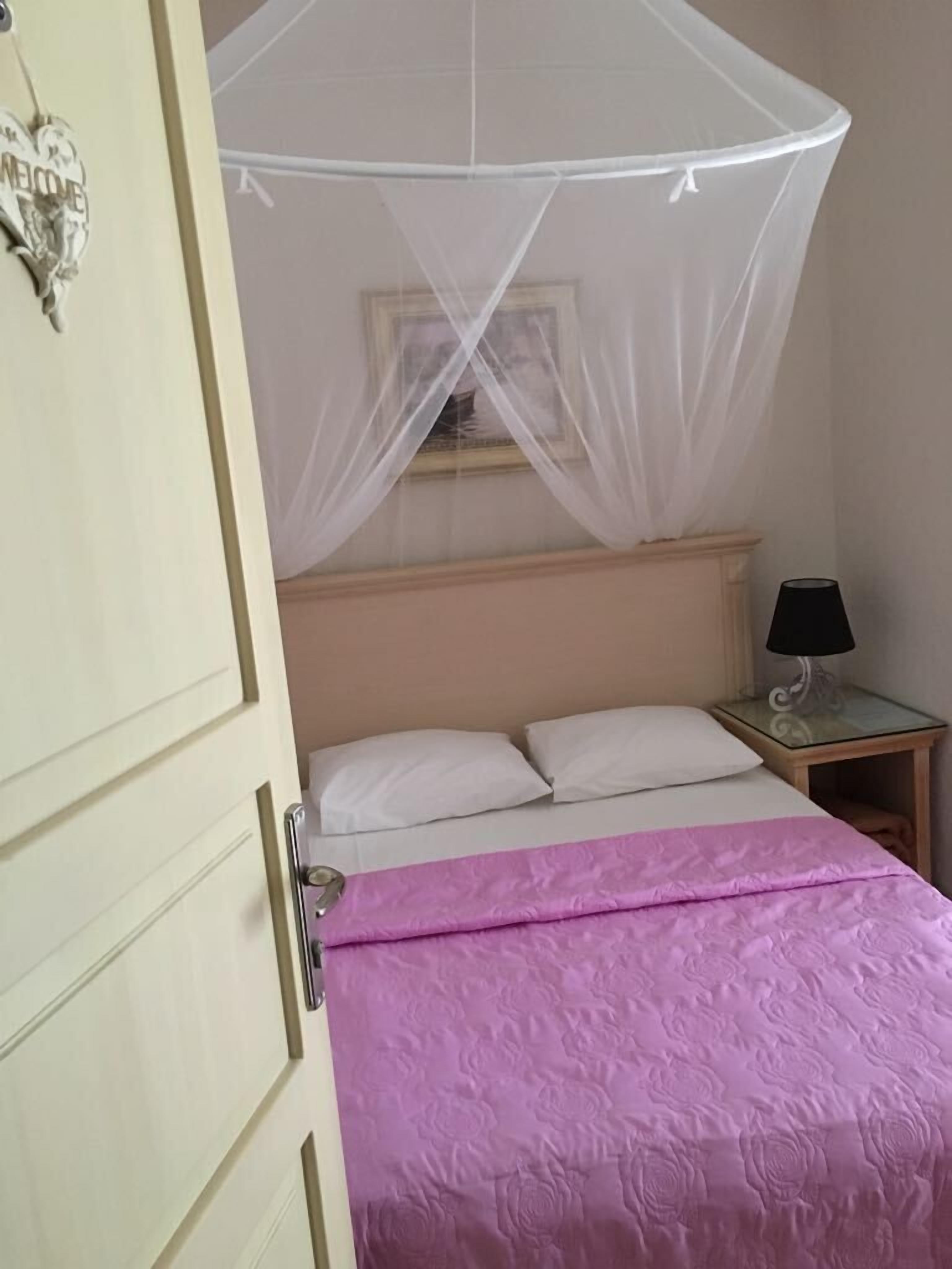 A bedroom with a bed under a mosquito net, a nightstand with a lamp, and a door with a 'WELCOME' sign.