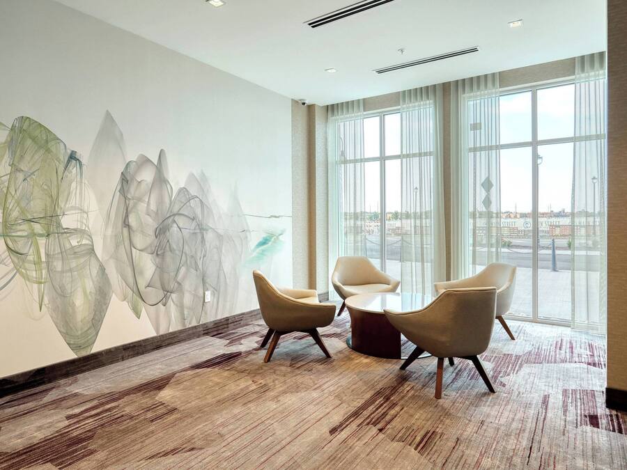 Un vestíbulo de hotel moderno con arte abstracto en la pared, ventanas amplias y una zona de estar con tres sillas y una mesa.