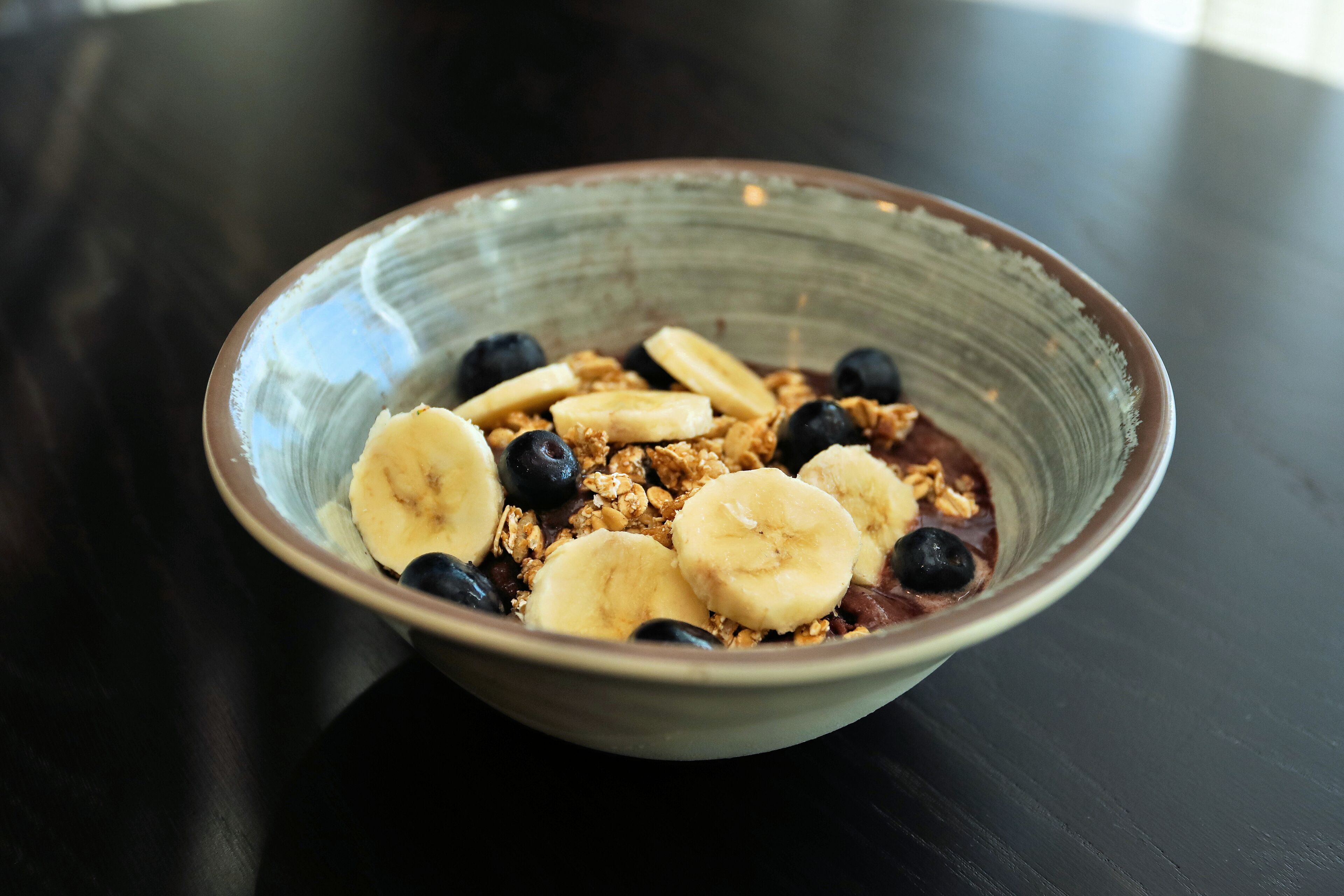 Un tazón de cereal con plátanos y arándanos sobre una mesa oscura.