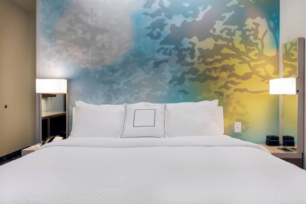 Habitación de hotel con cama, dos lámparas de noche, mesita de noche y una pared con estampado de camuflaje.