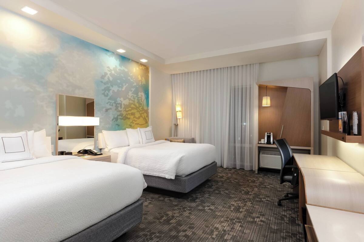 Habitación de hotel con dos camas, un escritorio, un televisor y un ventanal grande con cortinas.