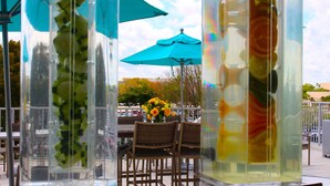 A glass-enclosed outdoor seating area with wicker chairs and a table, featuring a blue umbrella and a vase with yellow flowers. - DoubleTree by Hilton Miami - Doral, FL (Miami)