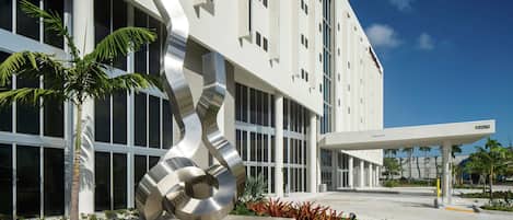 A modern building with a large metallic sculpture in front, a covered entrance, and a clear blue sky.