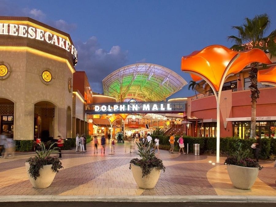 Un centro comercial con un letrero destacado que dice 'Dolphin Mall', una tienda 'Cheesecake' y una gran estructura en forma de cúpula iluminada.
