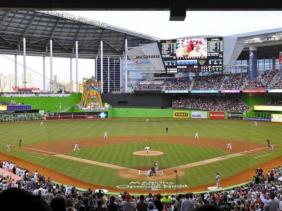 A baseball stadium with a large screen displaying game information, advertisements, and a mascot.