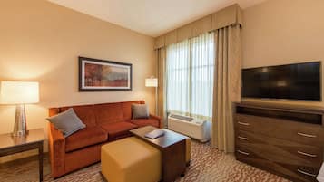A hotel room with a sofa, ottoman, flat-screen TV, air conditioning unit, and a lamp.