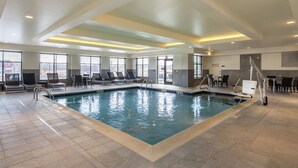 Una piscina interior con agua cristalina, rodeada de sillas de descanso y mesas, y amplias ventanas que ofrecen vistas al exterior.