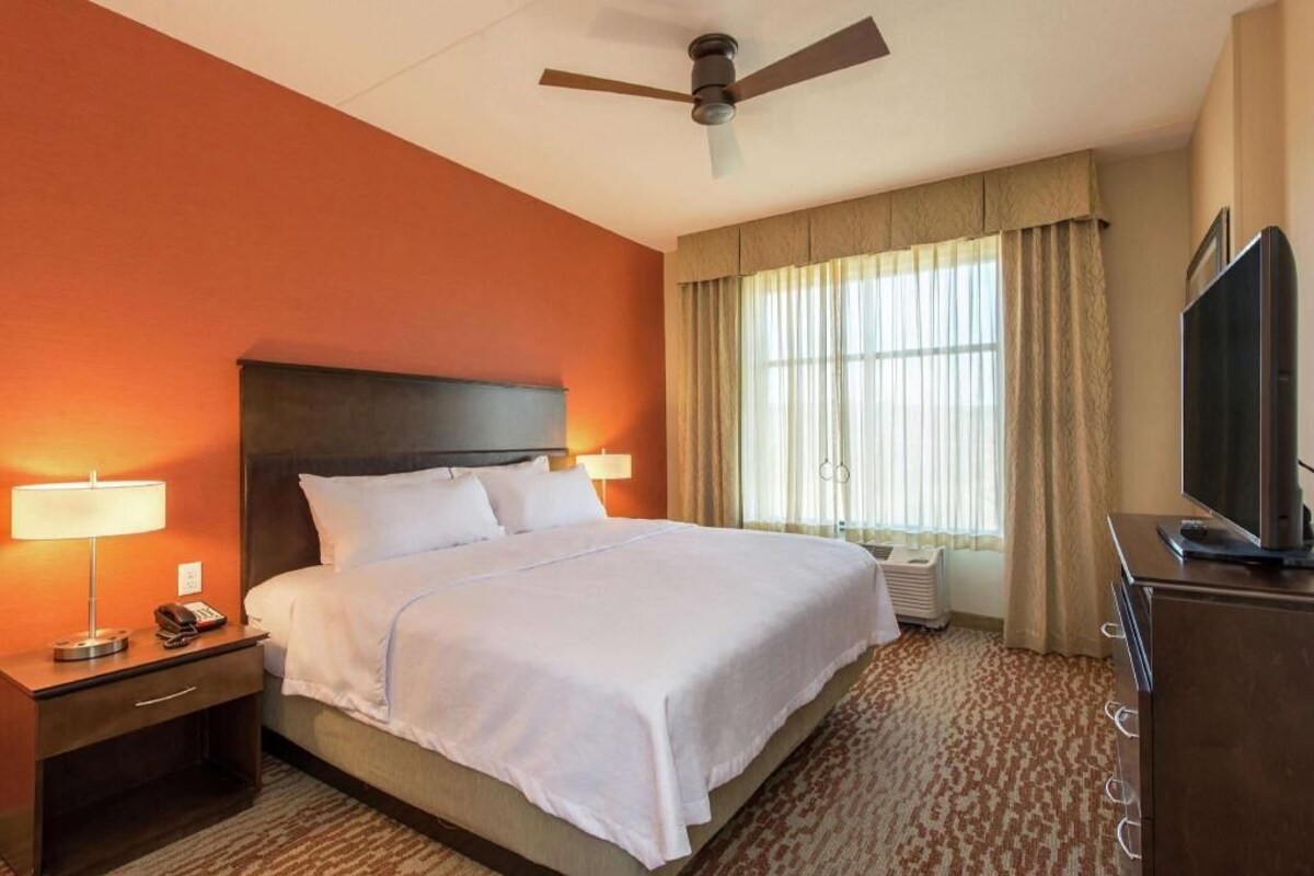 Una habitación de hotel con una cama, un escritorio con lámpara, un televisor y un ventilador de techo.
