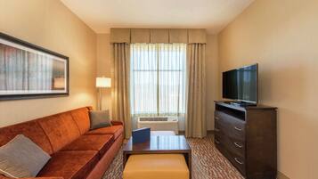 A hotel room with a brown sofa, a small table, a TV on a cabinet, and a window with curtains.