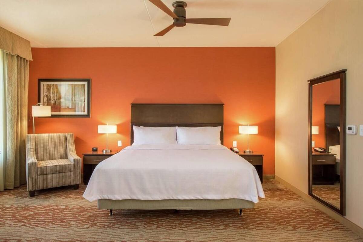 Een hotelkamer met een bed, een stoel, een nachtkastje, een lamp, een plafondventilator en een spiegel.