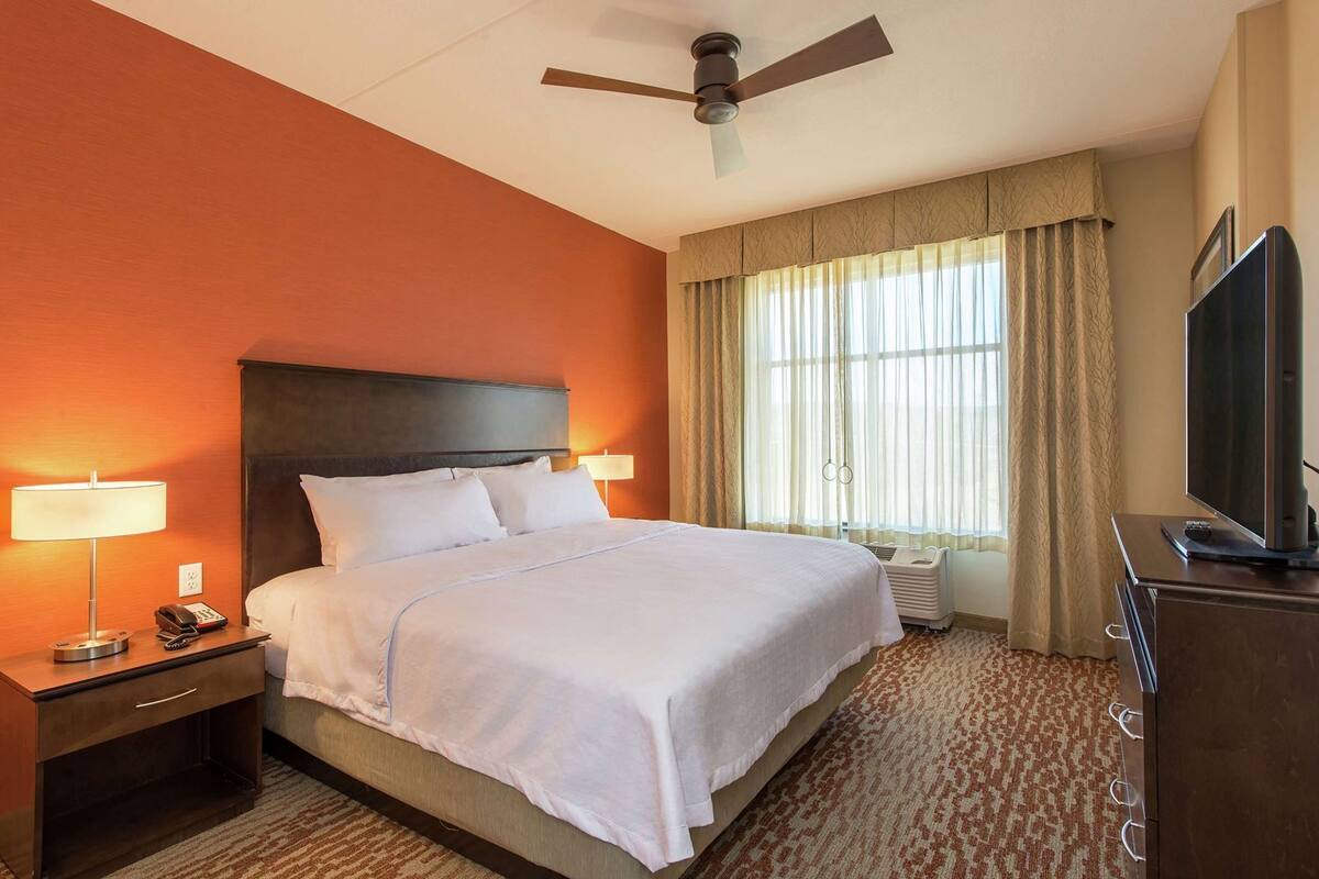 Een hotelkamer met een bed, bedlampen, een plafondventilator, een televisie en een raam met gordijnen.
