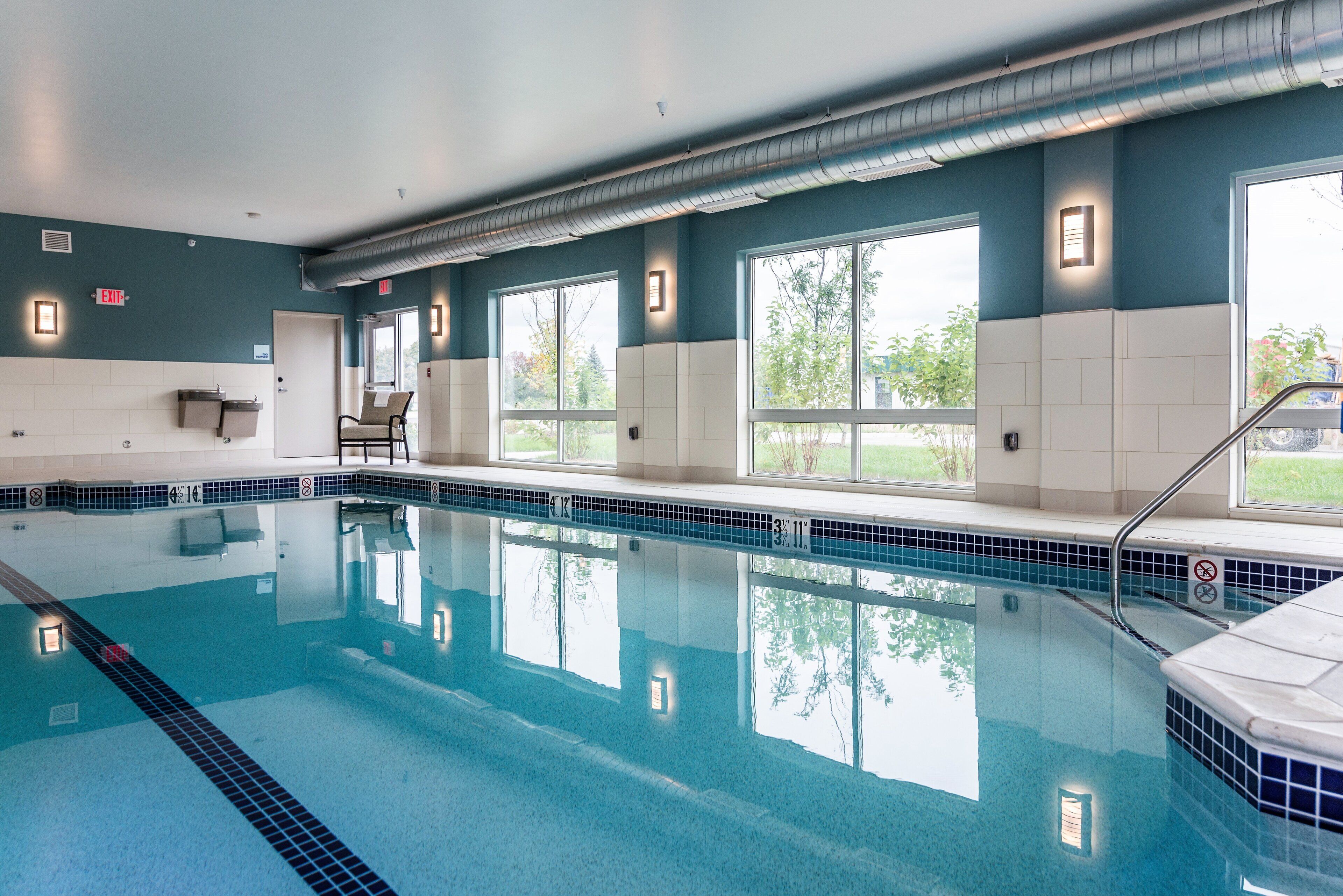 A swimming pool with clear water, surrounded by walls with windows, and an exit sign.