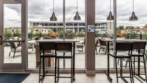 An indoor seating area with tables and chairs, large windows with a view of a construction site, and hanging pendant lights. - Holiday Inn Express & Suites Allen Park by IHG (Allen Park)