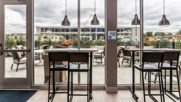 An indoor seating area with tables and chairs, large windows with a view of a construction site, and hanging pendant lights. - Holiday Inn Express & Suites Allen Park by IHG (Allen Park)