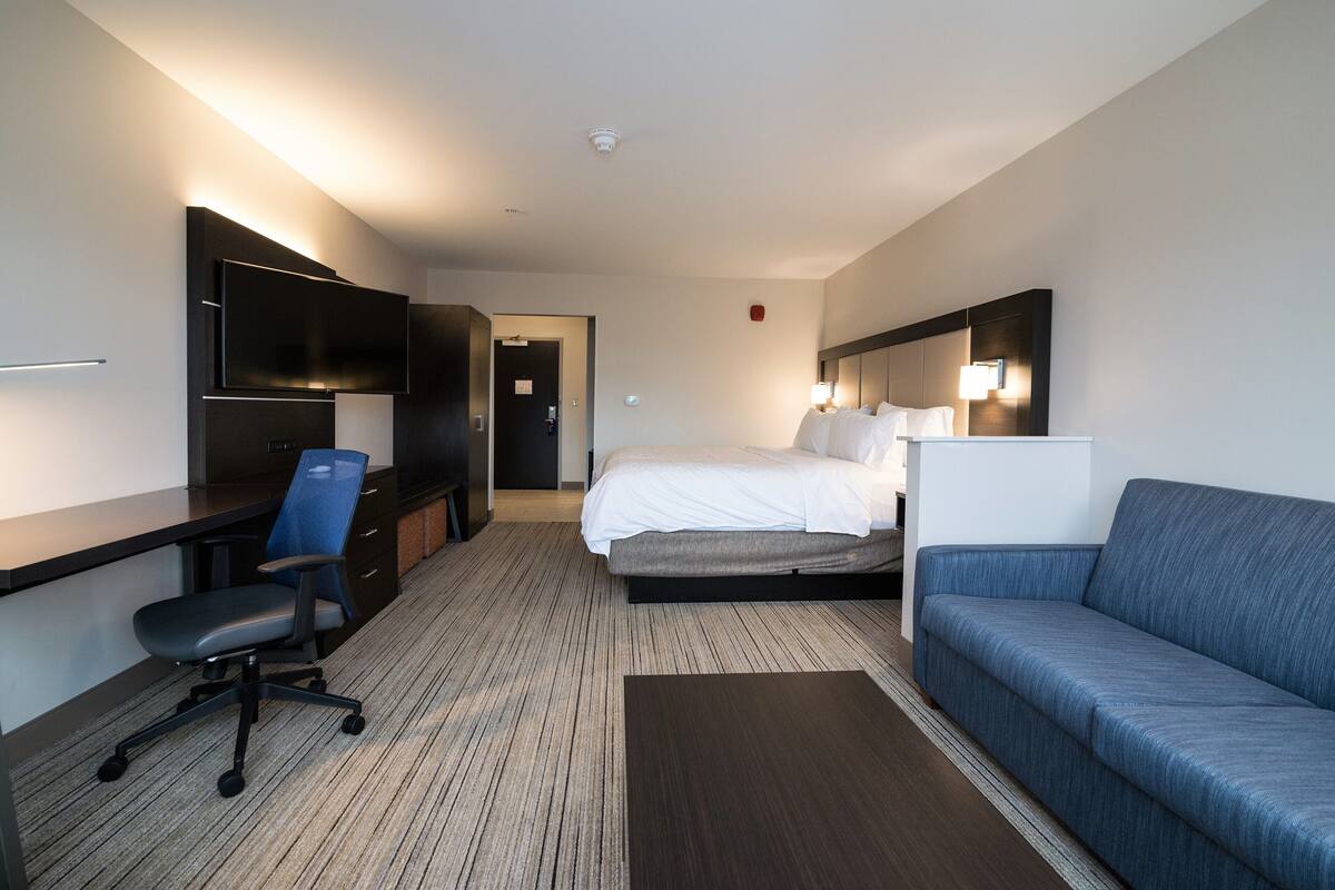 Una camera d'albergo moderna con un letto, una scrivania con sedia, un divano, una TV a schermo piatto e una lampada da comodino.