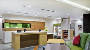 A modern hotel lobby with a reception desk, a seating area with a green sofa, a small table, and a refrigerator stocked with beverages. - Home2 Suites by Hilton Fort Worth Fossil Creek (Fort Worth)