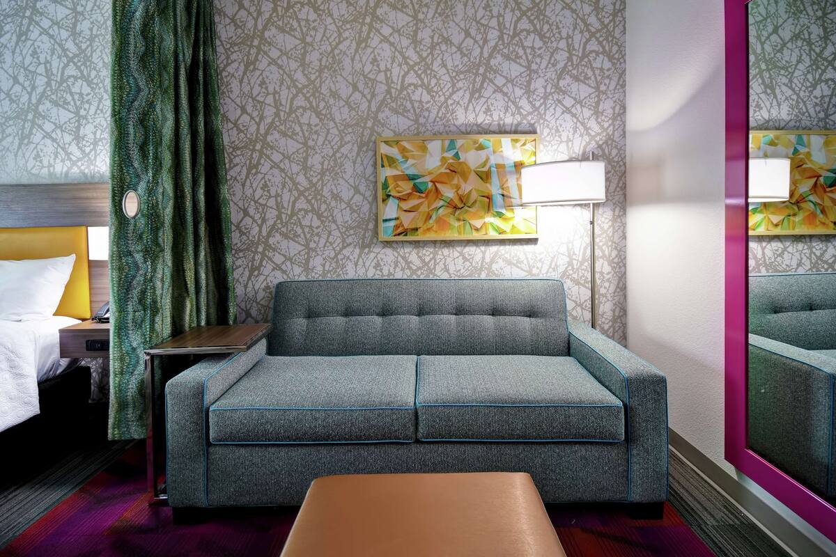 Een hotelkamer met een grijze bank, een klein tafeltje, een bed met witte lakens, een groen gordijn en een muur met behang met een geometrisch patroon.