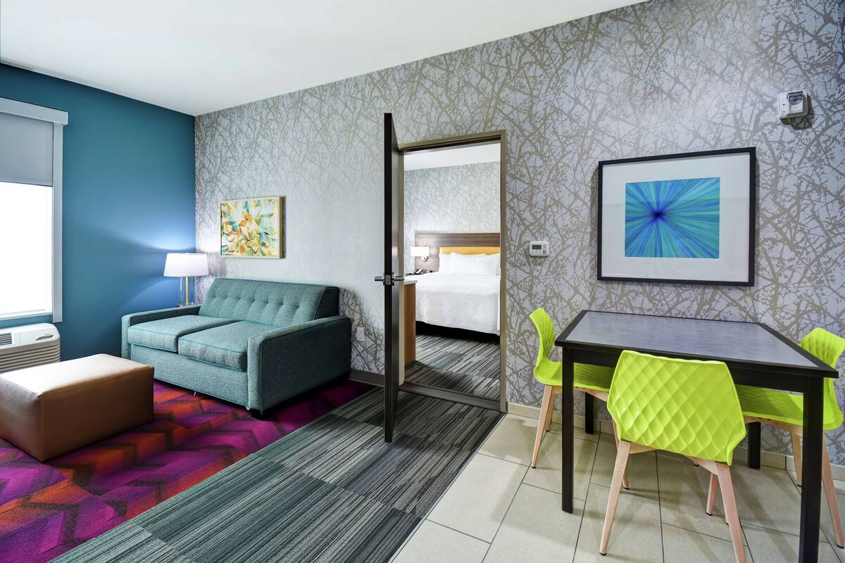 Een hotelkamer met een blauwgroene bank, een eettafel met groene stoelen, een kamerbreed tapijt en een lijst met abstracte kunst aan de muur.