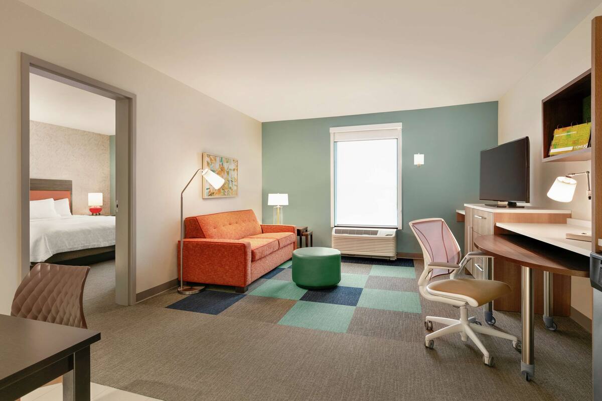 Een hotelkamer met een bed, een oranje sofa, een groene poef, een bureau, een stoel en een televisie.