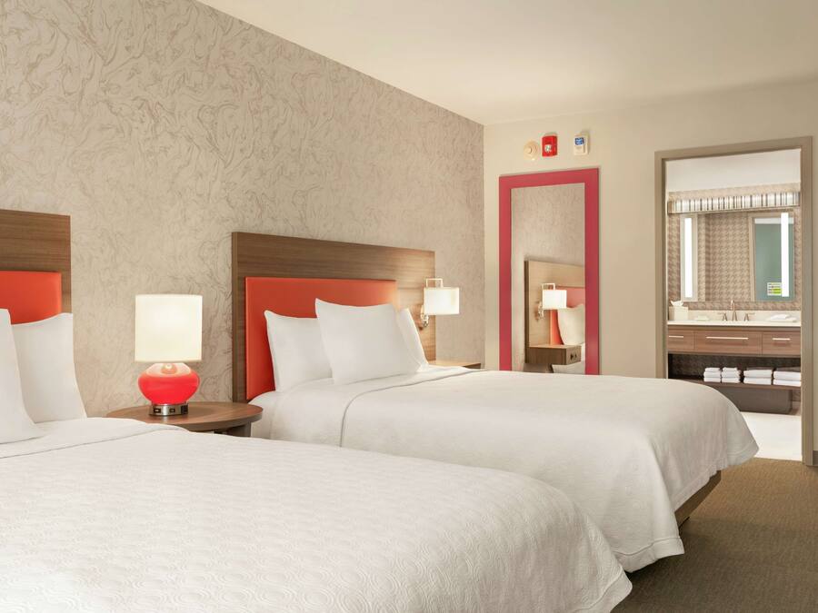 A hotel room with two beds, a red lamp, a mirror, and a bathroom visible through an open door.