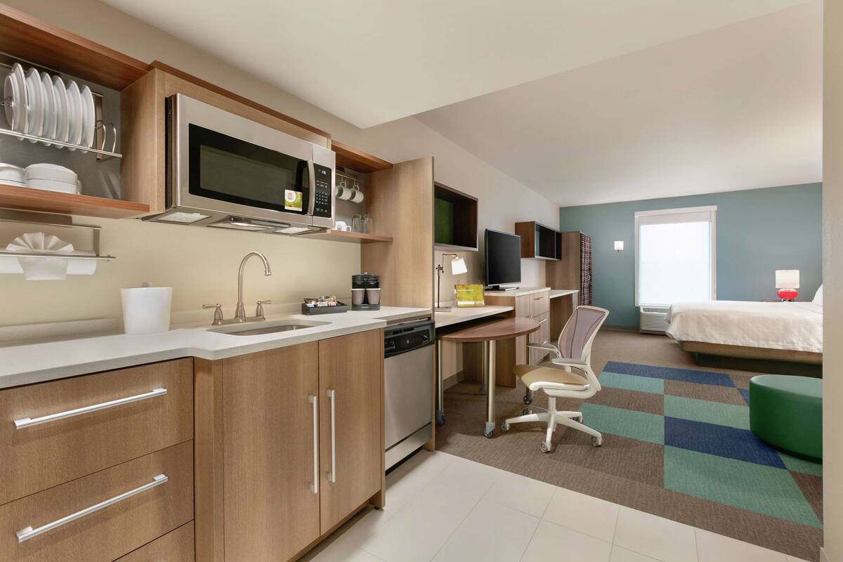 Een moderne keuken met een magnetron, spoelbak en vaatwasser. Er is een eethoek met een tafel en stoelen. Een bed is op de achtergrond zichtbaar.