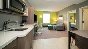 A modern kitchen with a built-in microwave, a sink, and a coffee maker. There is a dining area with a table and chairs, and a living room with a sofa and a lamp. - Home2 Suites by Hilton Jackson Flowood Airport Area (Flowood)