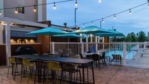 An outdoor patio with a long table and chairs, surrounded by umbrellas, and a pool in the background. - Home2 Suites by Hilton Jackson Flowood Airport Area (Flowood)