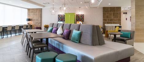 A modern hotel lobby with a large seating area, a reception desk, and a dining area.