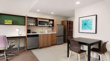 A modern kitchen with a microwave, refrigerator, and wooden cabinets. There is a dining table with chairs and a desk with a chair.