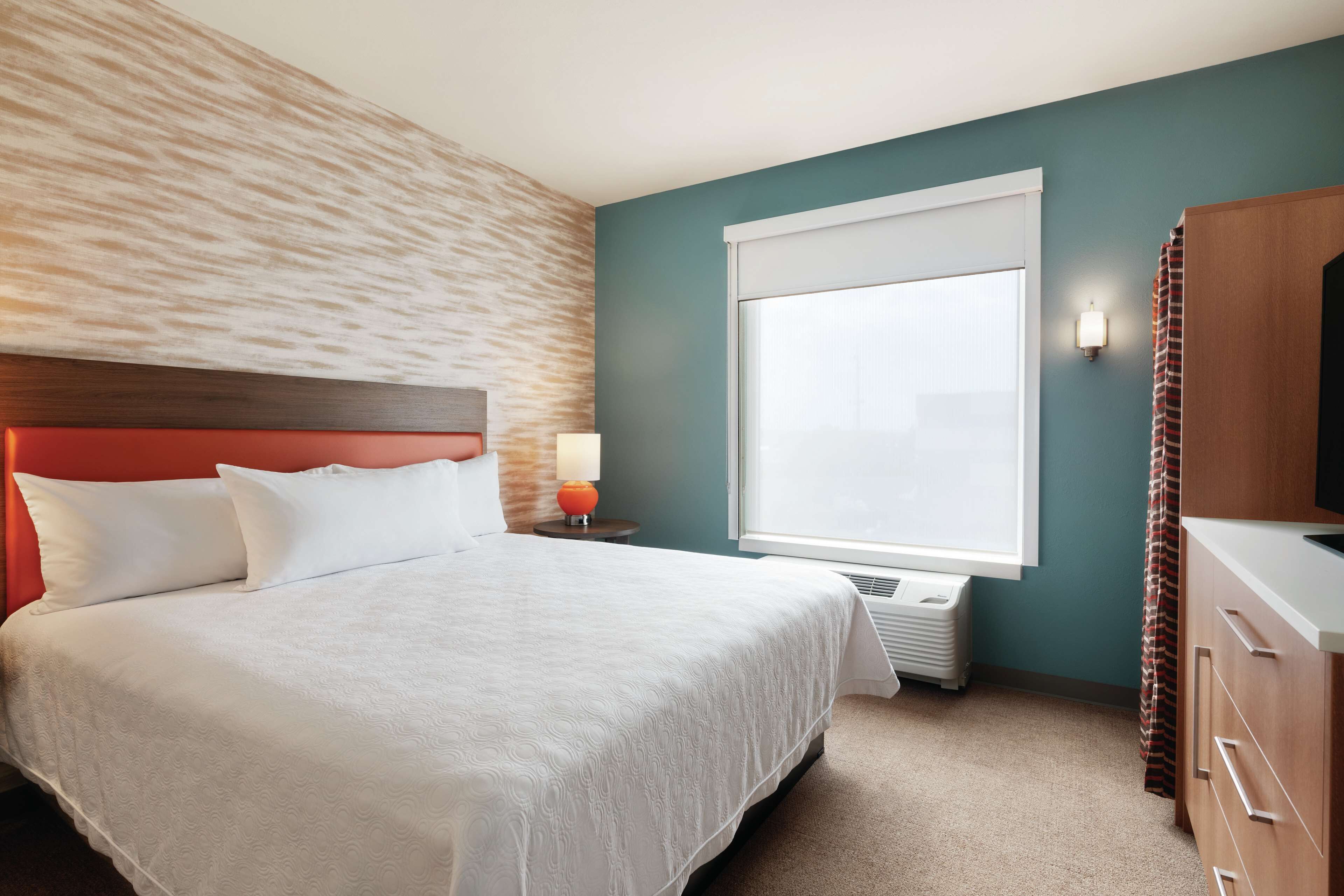 A hotel room with a large bed, a nightstand with a lamp, a window with blinds, a heater, and a wooden dresser.