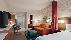 Una habitación de hotel con cama, escritorio con computadora, silla, sofá, televisión y baño visible a través de una puerta abierta.