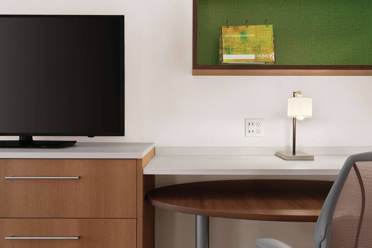 A modern hotel room with a flat-screen TV, a wooden desk, a lamp, and a bulletin board with notes.