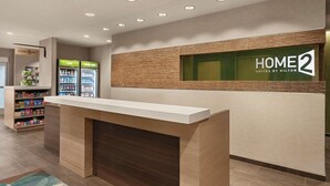Zona de recepción con mostrador, un letrero en la pared que dice "HOME 2 Suites by Hilton" y un refrigerador con bebidas.