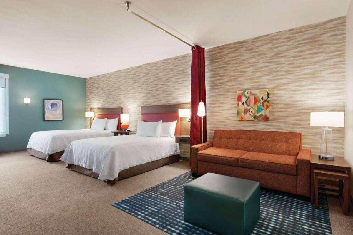 A hotel room with two beds, a sofa, a coffee table, a patterned rug, and a wall art piece.