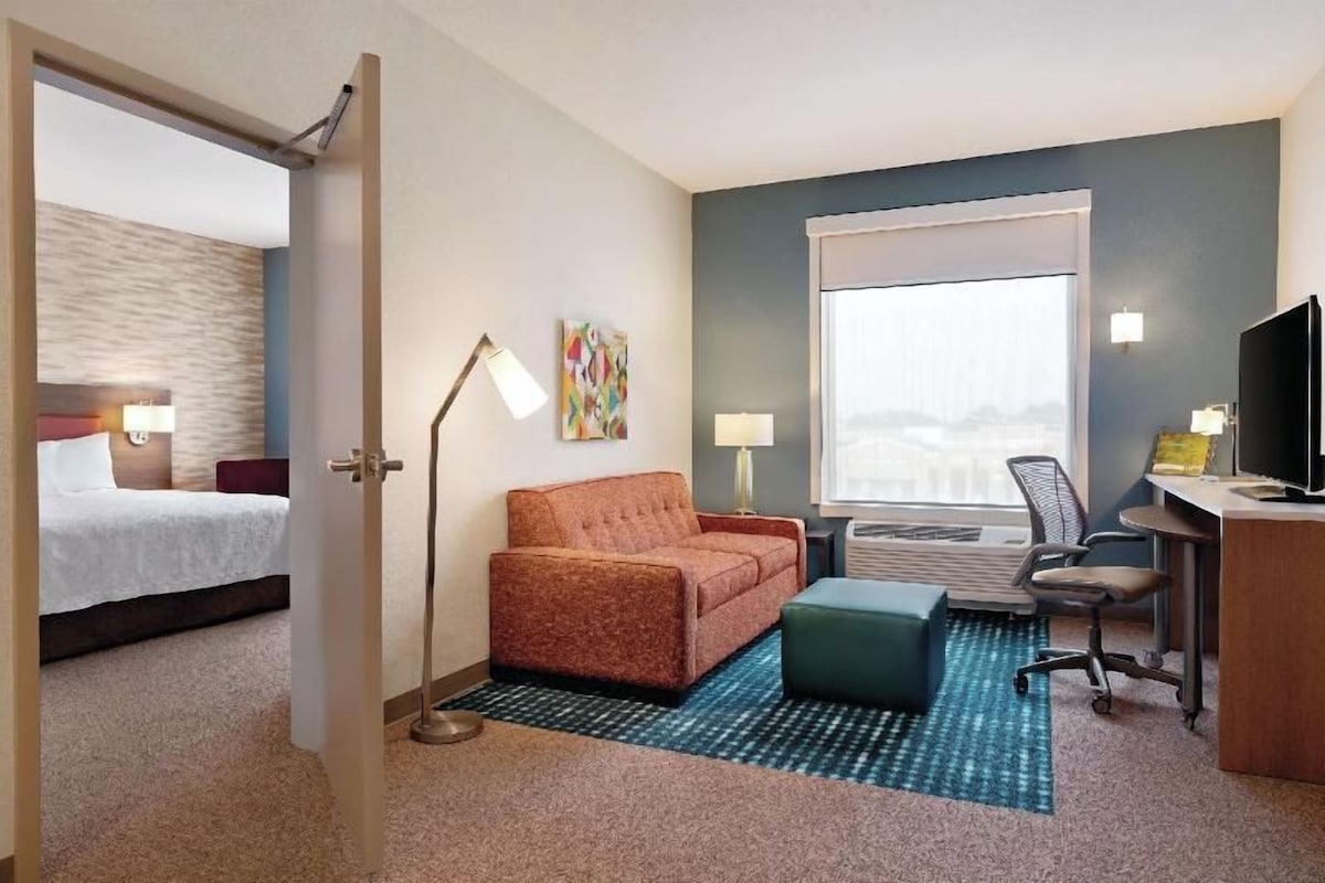 A hotel room with a bed, a sofa, a desk, a chair, a lamp, and a large window.