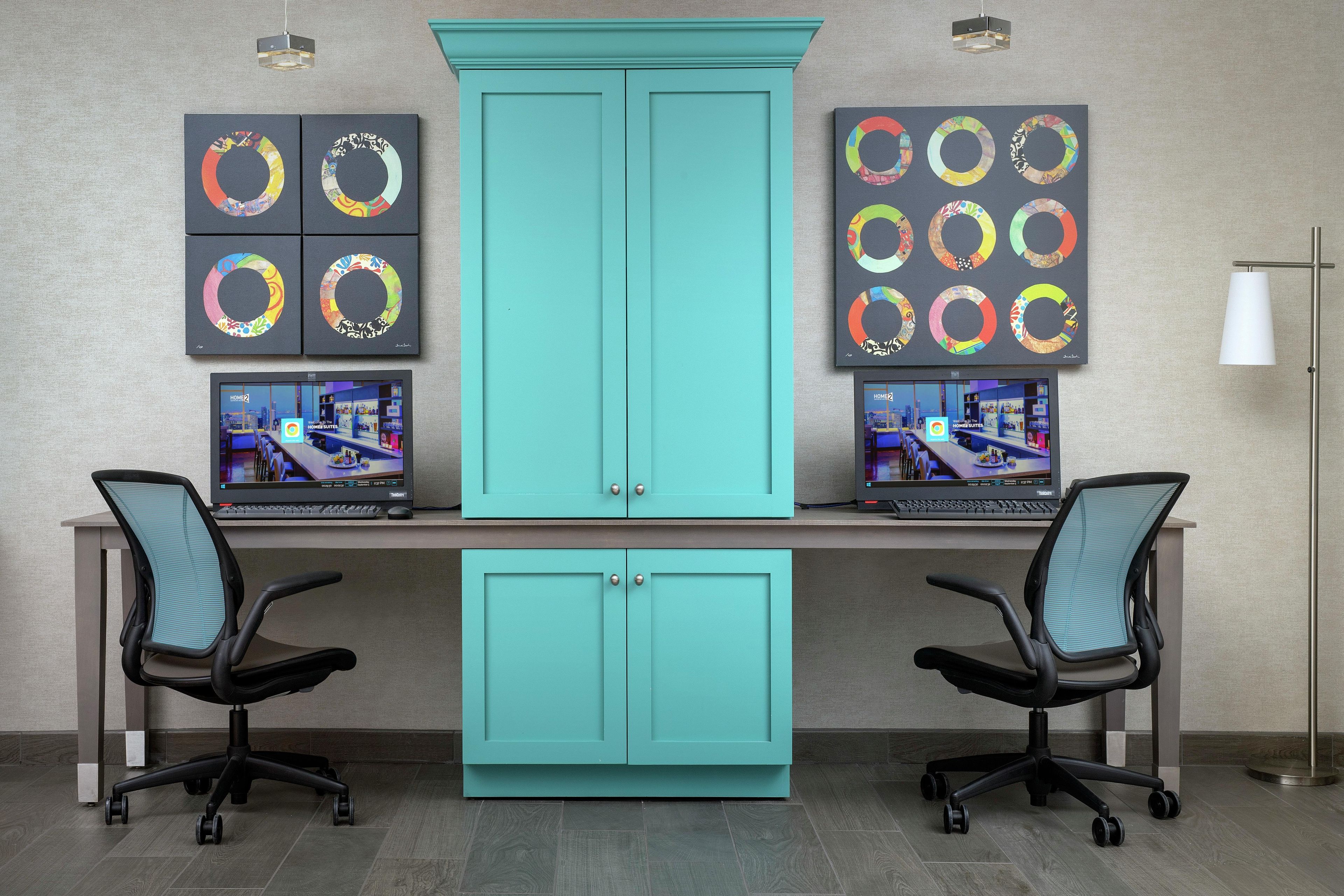 A teal-colored office desk with two monitors, two office chairs, and a cabinet.