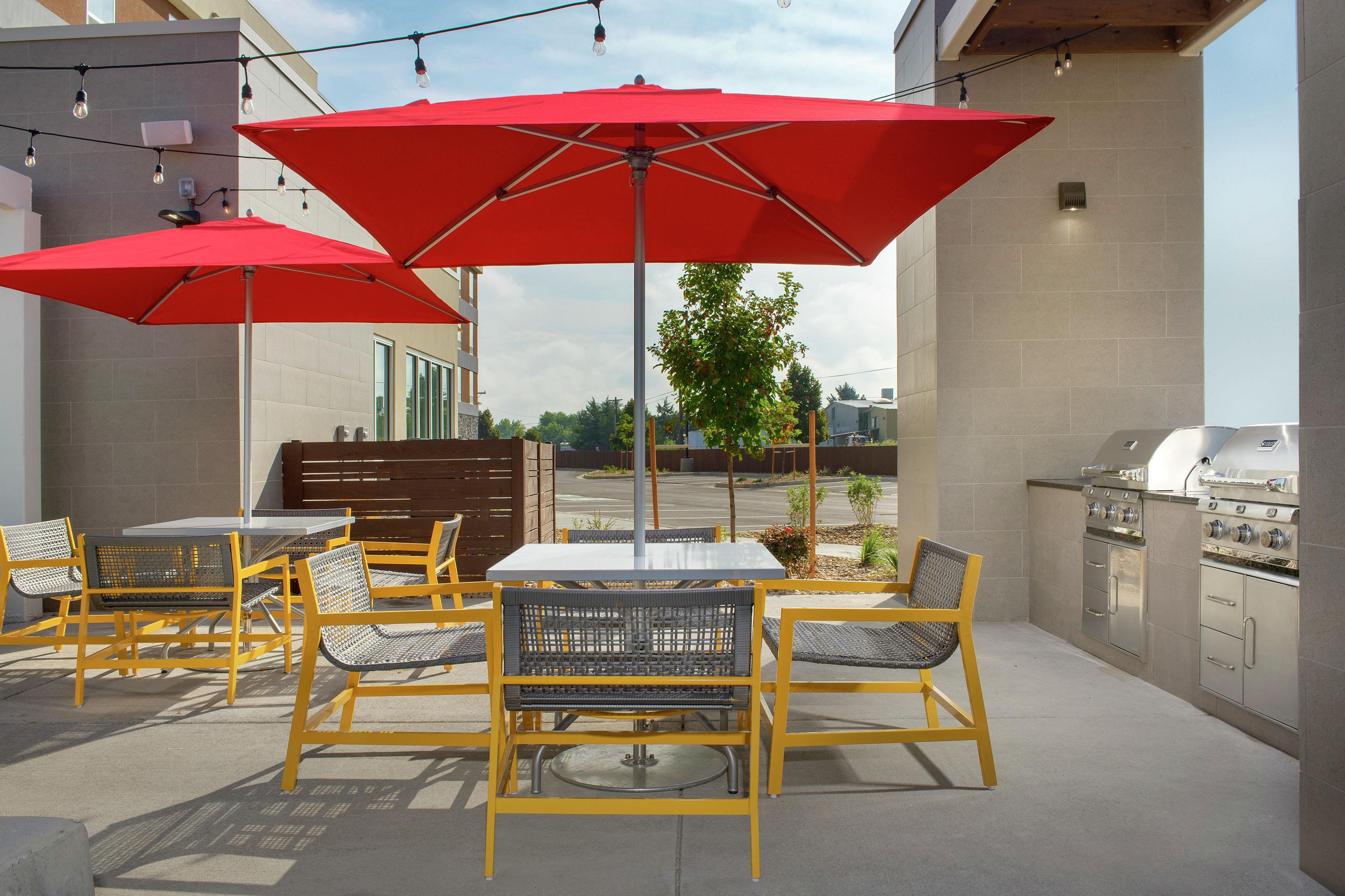 Un patio exterior con sillas amarillas y sombrillas rojas, una parrilla de barbacoa y vistas de la ciudad.
