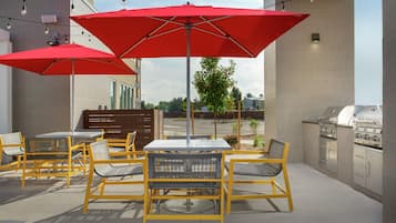 Un patio exterior con sillas amarillas y sombrillas rojas, una parrilla de barbacoa y vistas de la ciudad.