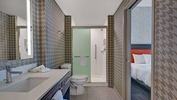Une salle de bain moderne avec un lavabo, des toilettes et une douche. Un miroir est placé au-dessus du lavabo et un porte-serviettes se trouve à côté. La salle de bain est décorée d’un papier peint à motifs et une porte donne sur une chambre à coucher où se trouve un lit.