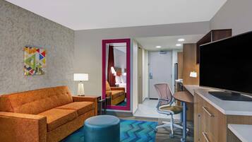 Quarto de hotel com sofá, apoio para os pés, escrivaninha, cadeira, televisão e espelho com moldura rosa.